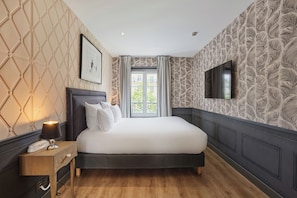A bedroom with a bed, a nightstand, a lamp, a framed picture, a window with curtains, and a wall-mounted TV. -  La Parizienne (Paris)