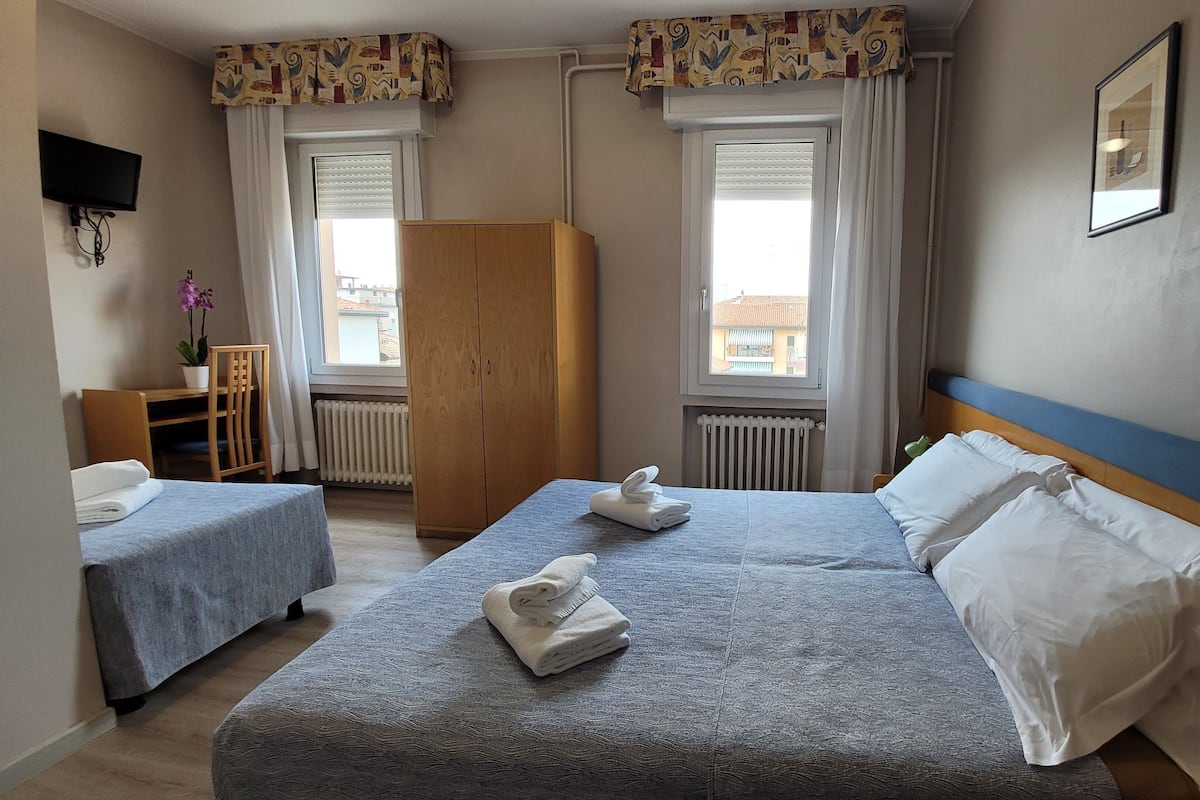 A hotel room with two beds, a wooden wardrobe, a desk with a chair, a radiator, and a TV mounted on the wall.
