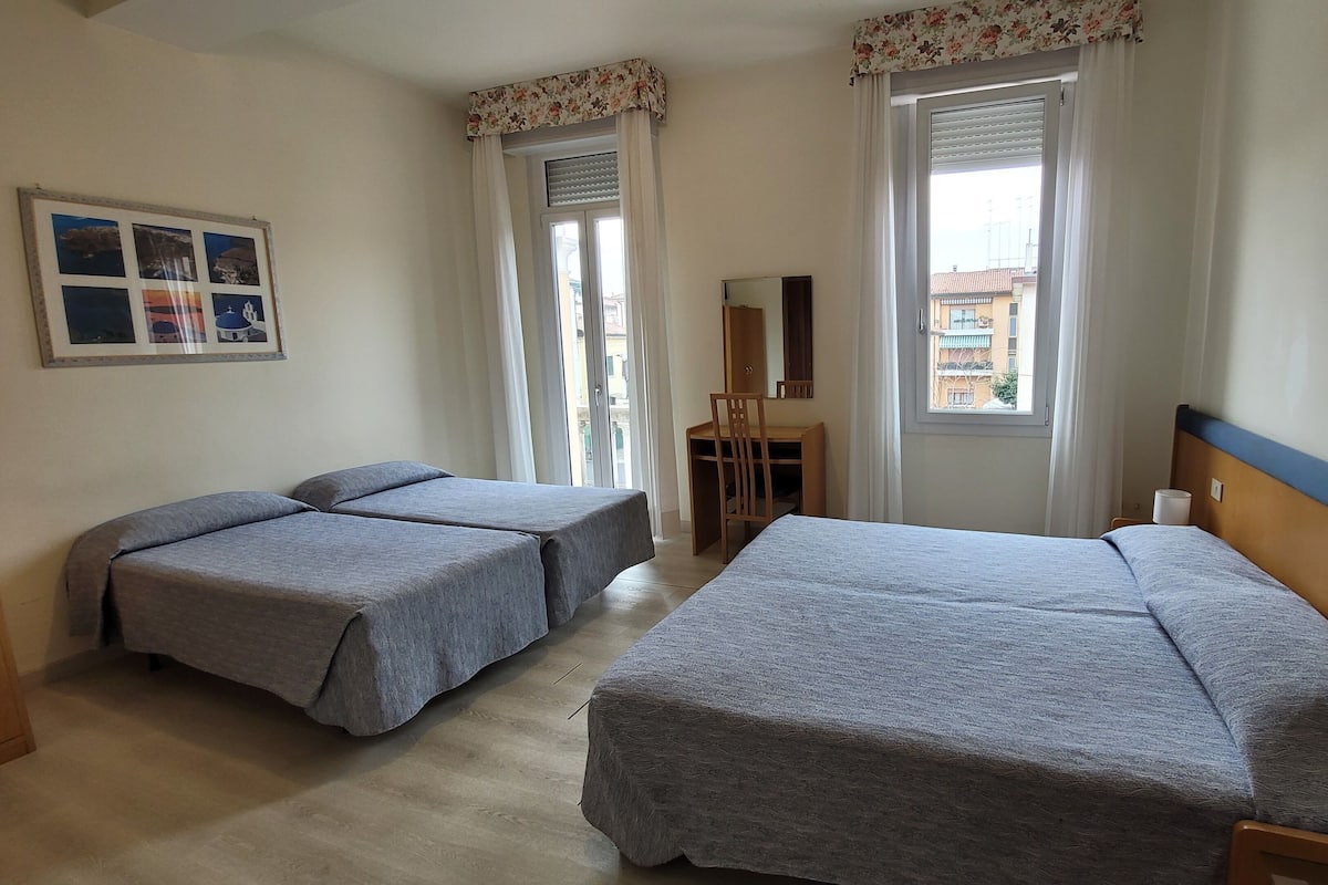 A hotel room with two single beds, a desk with a chair, a mirror, and a window with curtains.