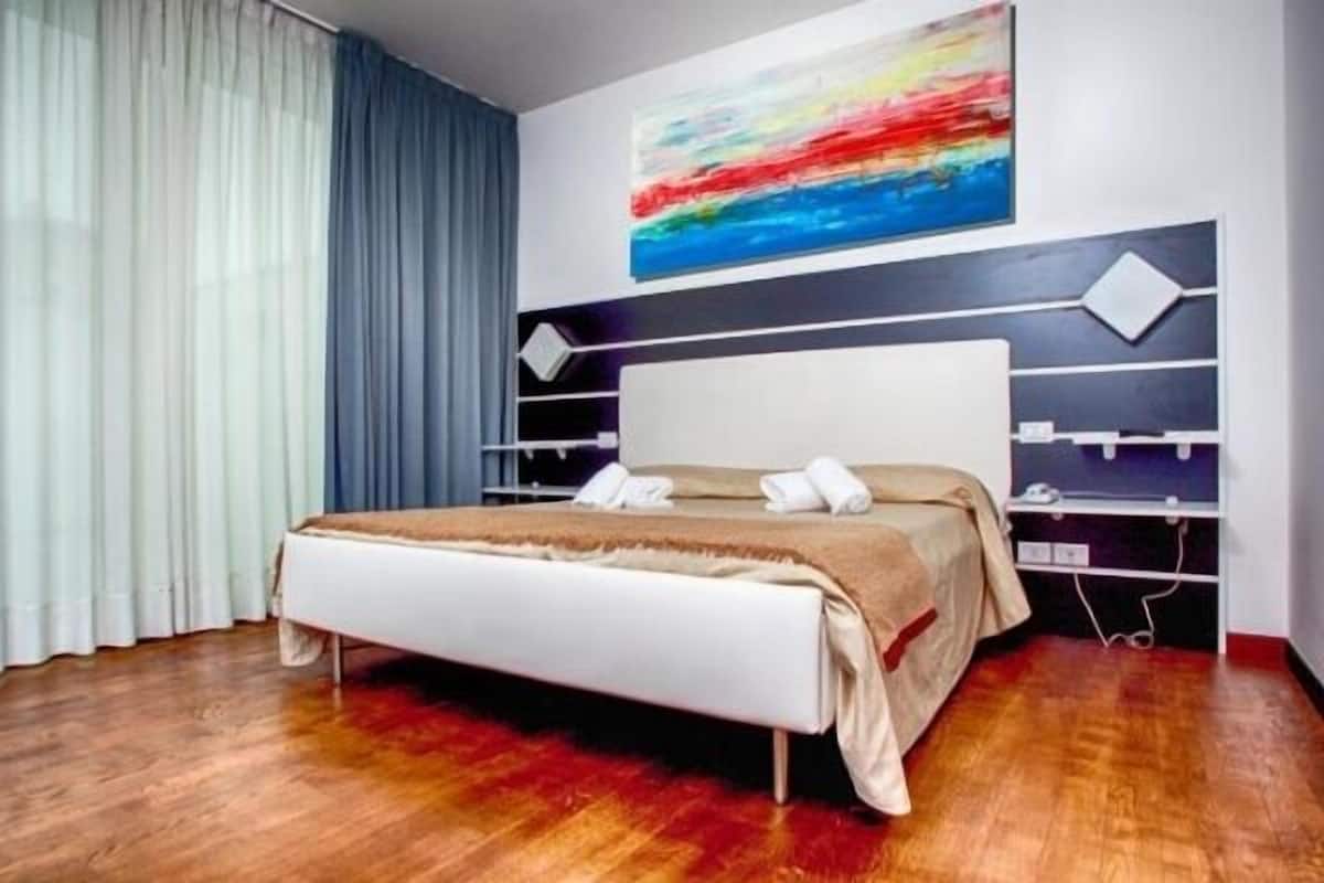 Una habitación de hotel moderna con una cama grande, un cabecero con espacio de almacenamiento y un cuadro abstracto de colores en la pared.