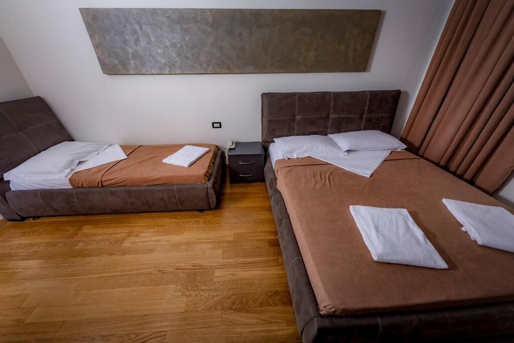 A hotel room with two beds, a wooden floor, a nightstand, and a painting on the wall.
