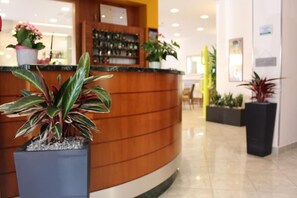 A reception area with a curved wooden counter, potted plants, and a tiled floor. - Angelo (Caorle)