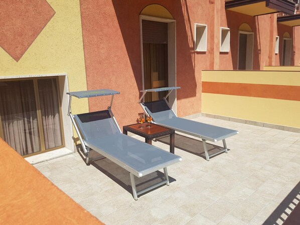 Terraza en la azotea con dos tumbonas y una pequeña mesa, con suelo de baldosas y un edificio de fachada amarilla y anaranjada.