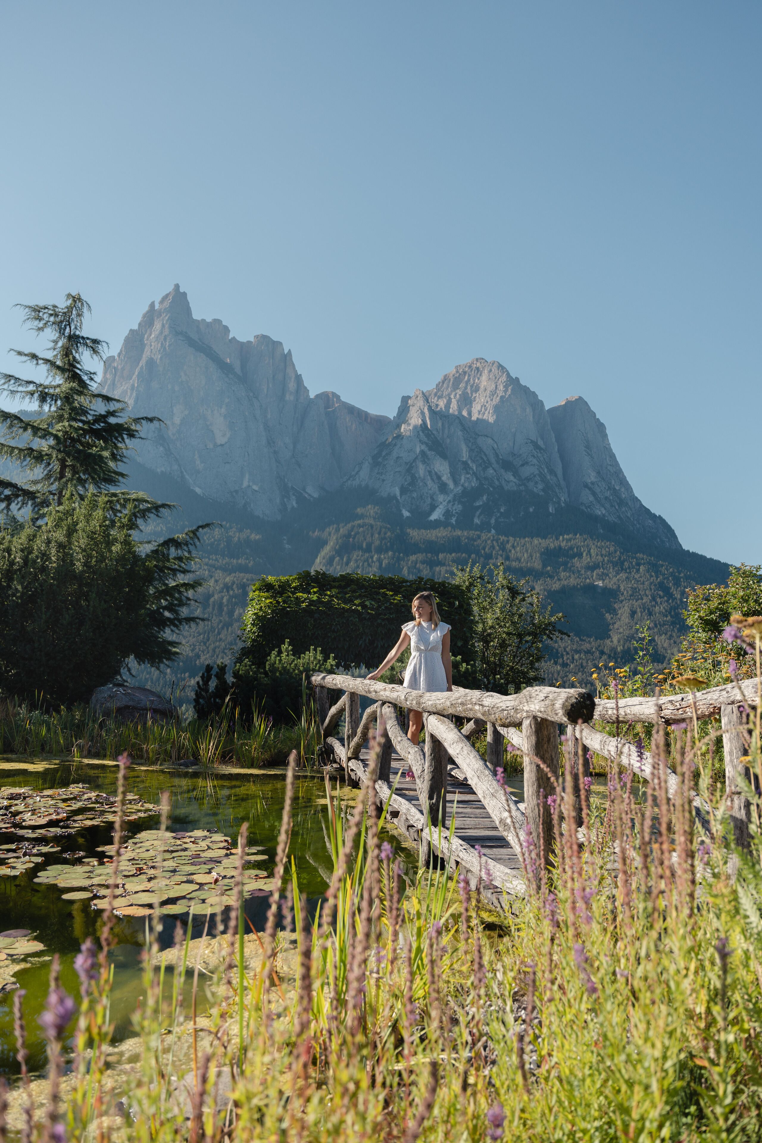 A woman in a white dress standing on a wooden bridge over a pond with lily pads, surrounded by lush greenery and mountains in the background.