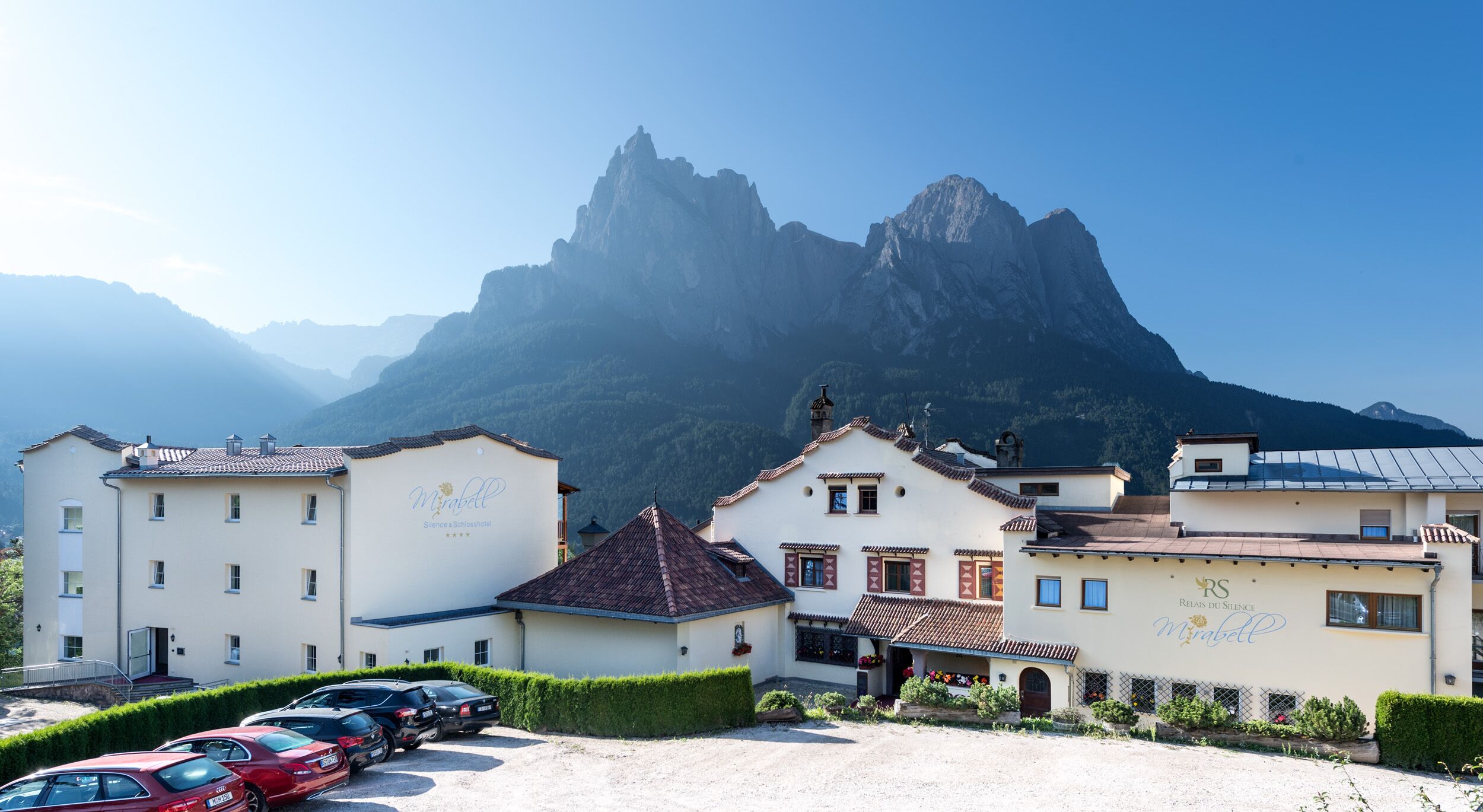 A hotel with a mountain backdrop, featuring a parking area with several cars.
