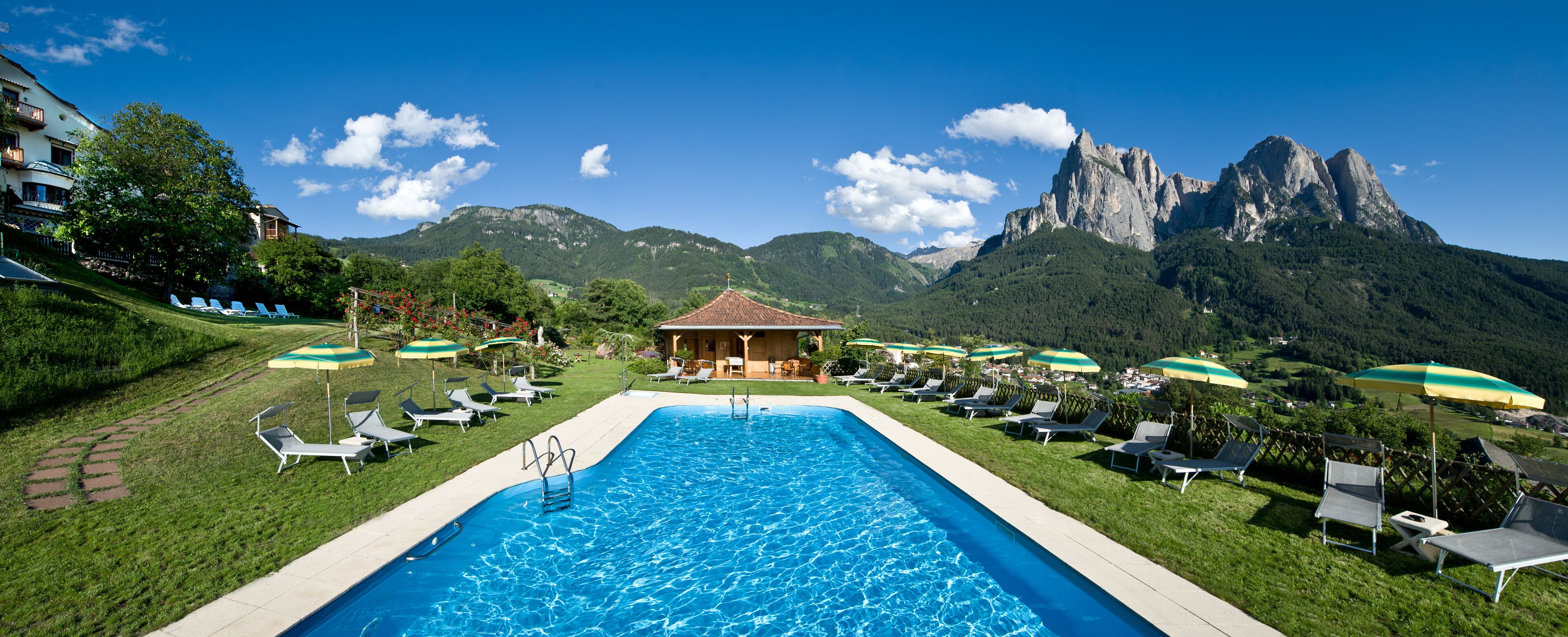 A swimming pool with lounge chairs and umbrellas, surrounded by mountains and a small building.