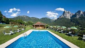 A swimming pool with lounge chairs and umbrellas, surrounded by mountains and a small building.