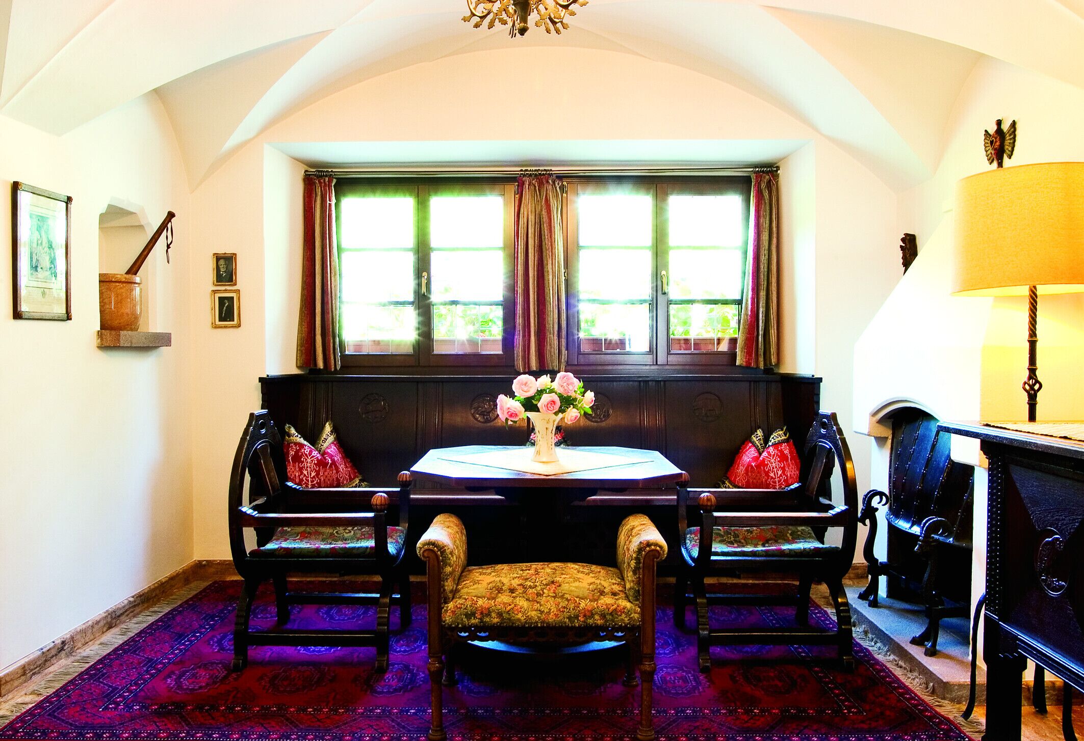 A room with a round table, two chairs, a bench, a window with curtains, a chandelier, and a lamp.