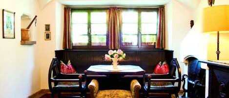 A room with a round table, two chairs, a bench, a window with curtains, a chandelier, and a lamp.