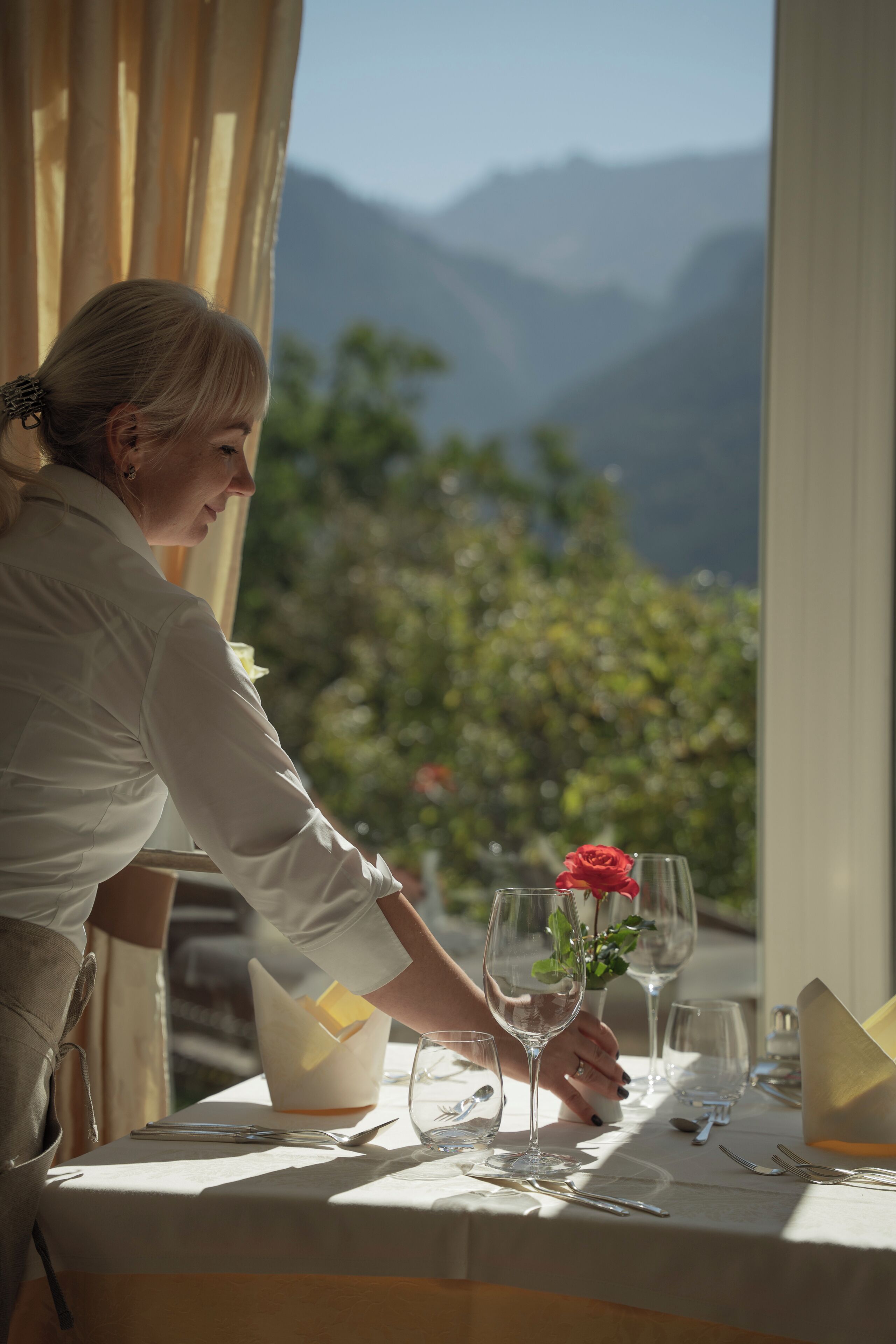 A dining table set with a wine glass and a red rose, with a mountainous landscape visible through a window.