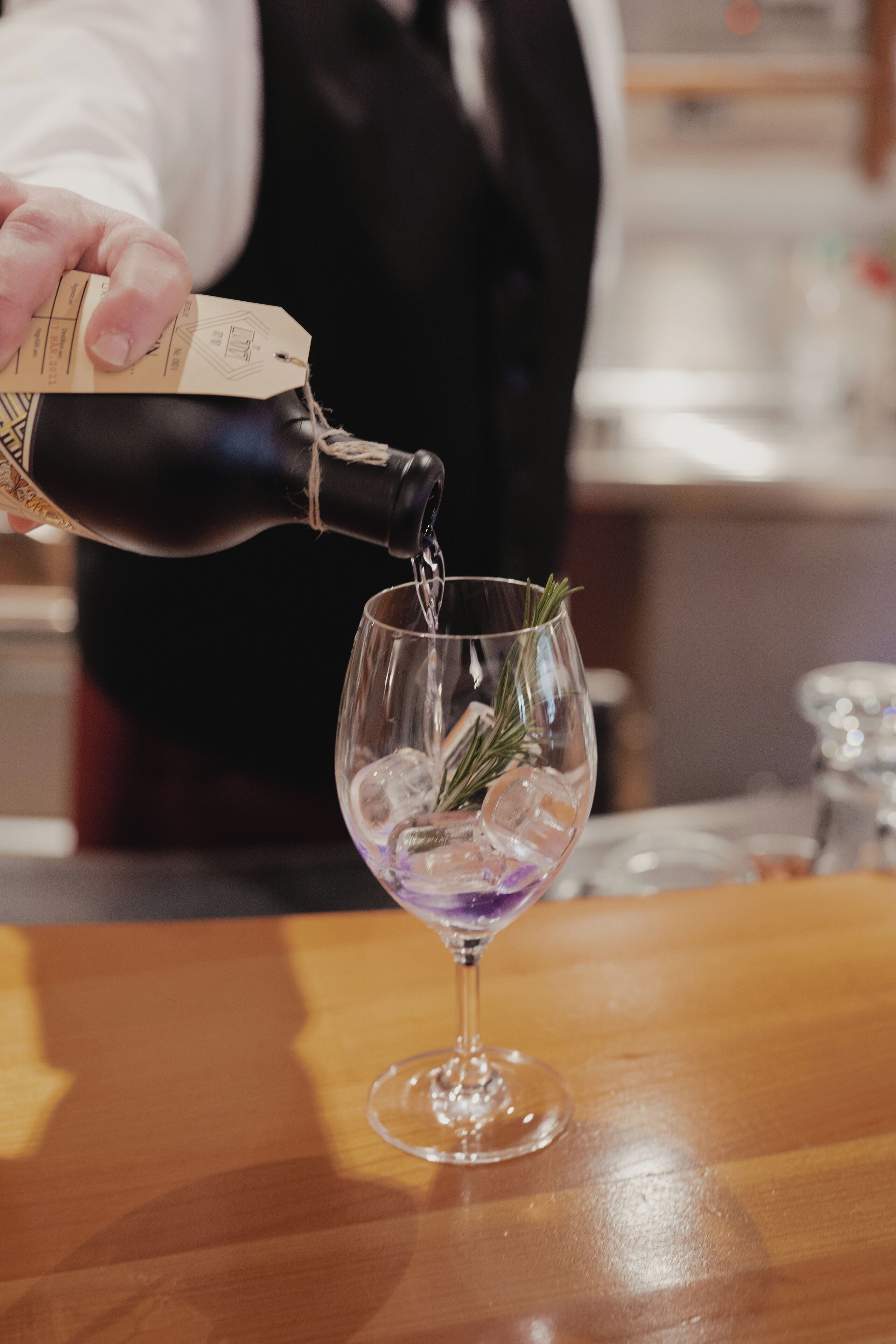 A person pouring a drink from a bottle into a glass with ice and a sprig of rosemary.