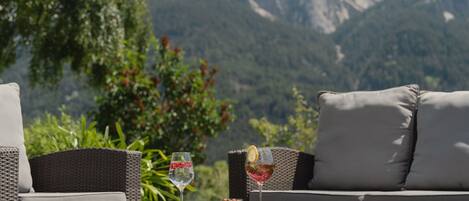 A wicker outdoor seating area with a glass table, two cushions, and a glass of wine.