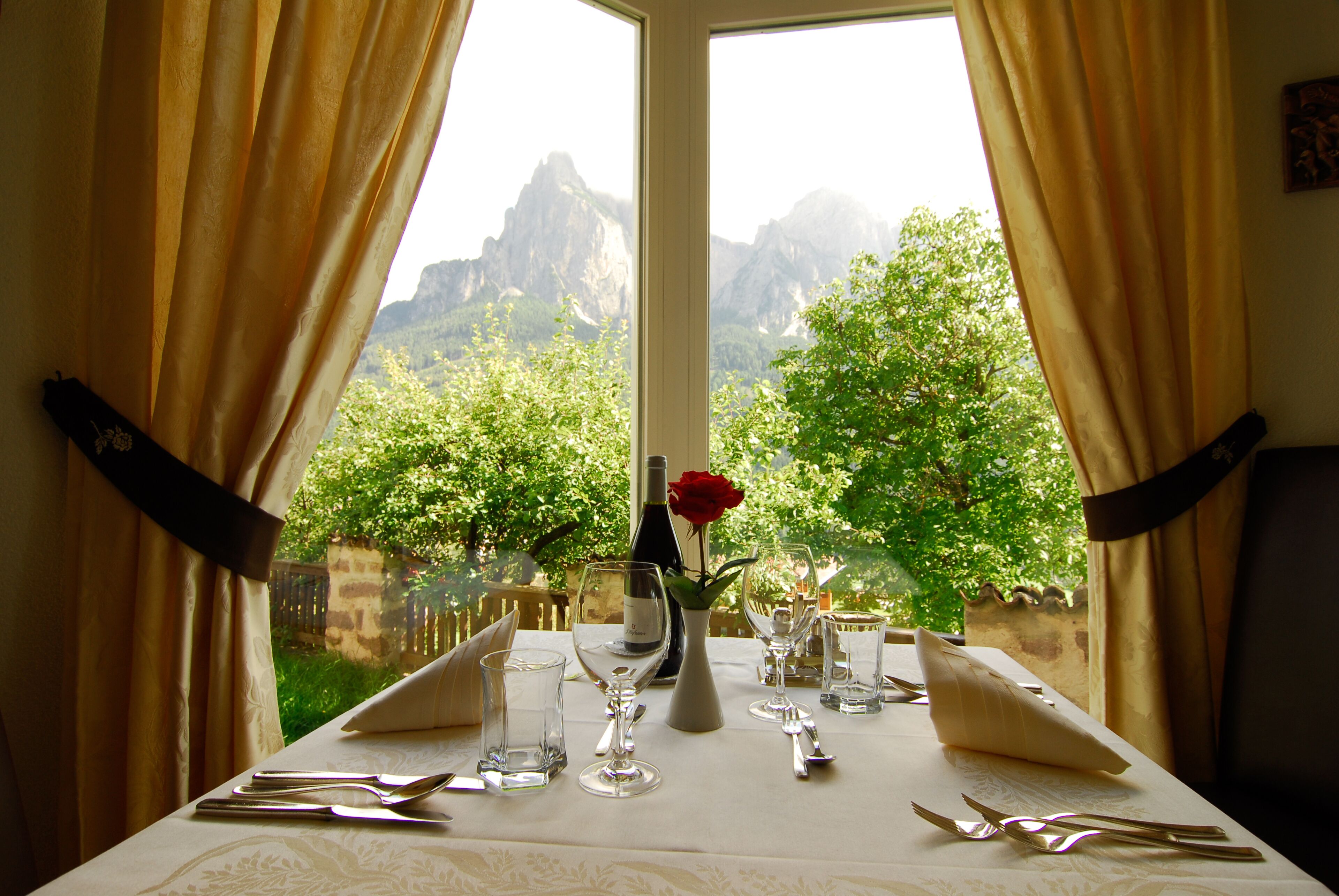 Una mesa de comedor dispuesta con cubiertos y vasos, un florero con una rosa roja y una botella, con vista a la montaña a través de una ventana.