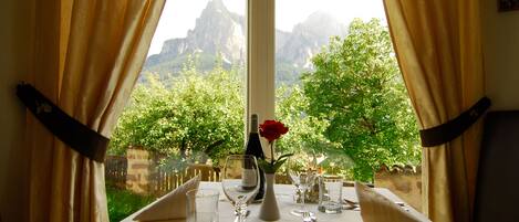Una mesa de comedor dispuesta con cubiertos y vasos, un florero con una rosa roja y una botella, con vista a la montaña a través de una ventana.