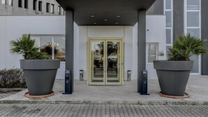 La entrada de B&B Hotels con un diseño moderno, que incluye un porche cubierto, plantas grandes en macetas y un amplio ventanal.
