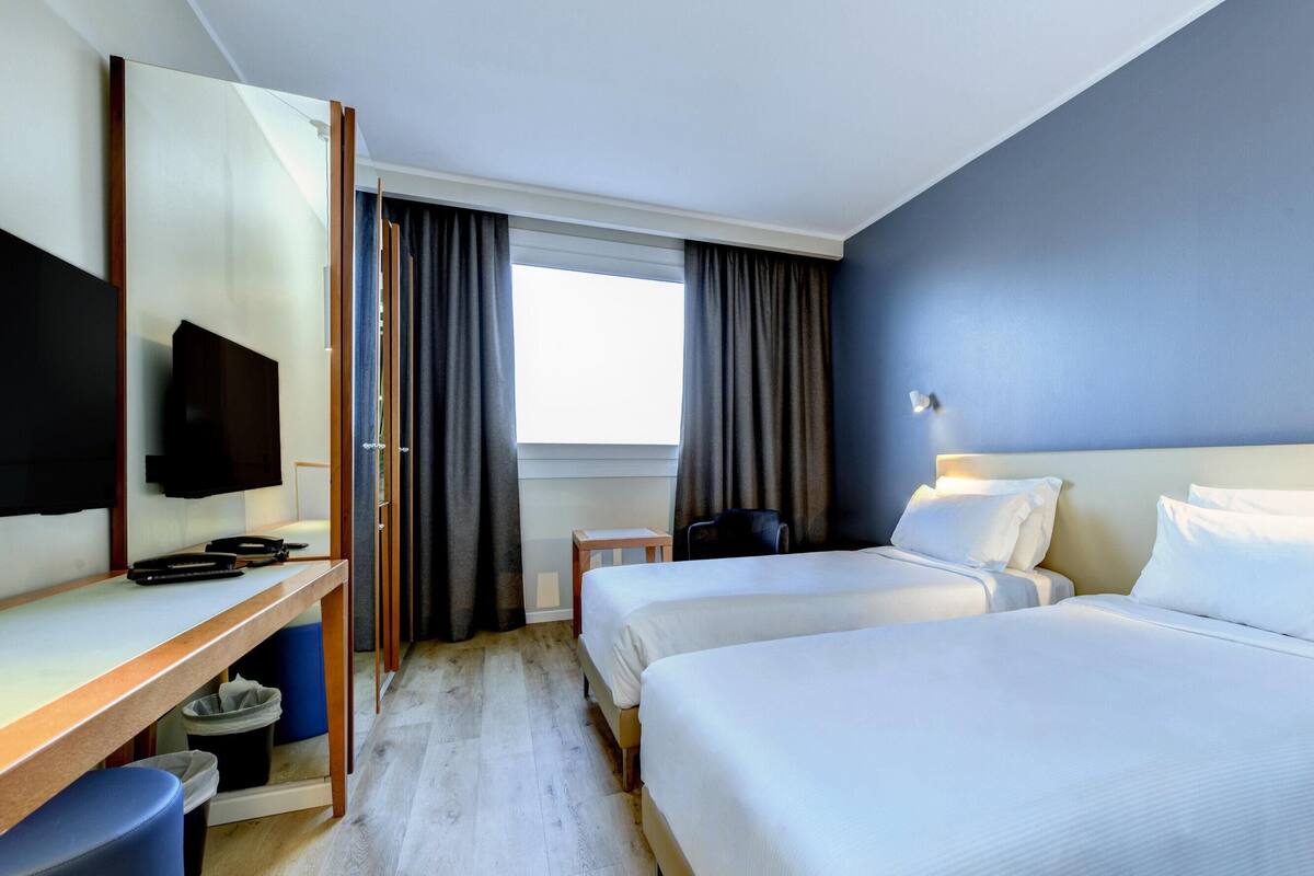A hotel room with two beds, a desk with a flat-screen TV, a chair, and a window with curtains.