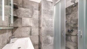 Un baño moderno con lavamanos blanco, cabina de ducha con mampara de vidrio y baldosas de piedra gris.