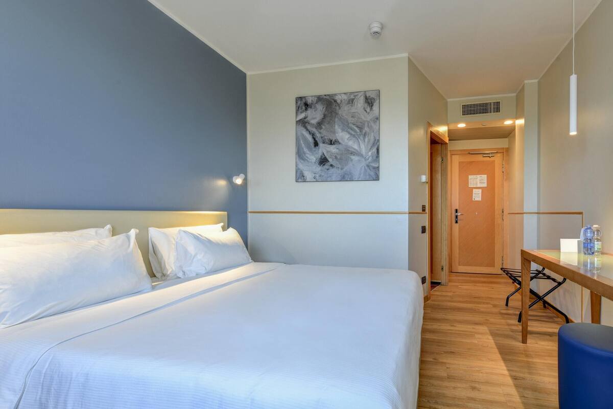 A hotel room with a large bed, a desk with a chair, a blue ottoman, and a painting on the wall.