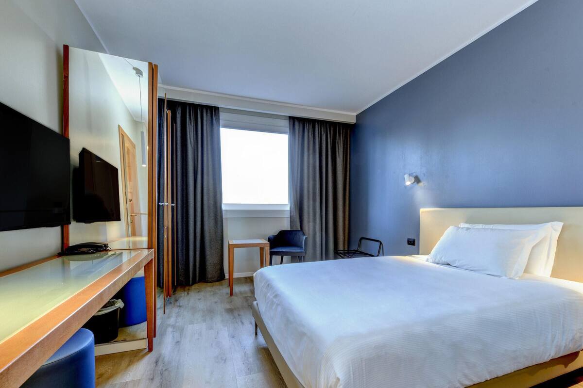 A hotel room with a bed, a desk, a chair, a TV, and a window with curtains.