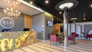 Zona de recepción de un hotel con mostrador de mármol, asientos rojos y una lámpara de araña moderna.