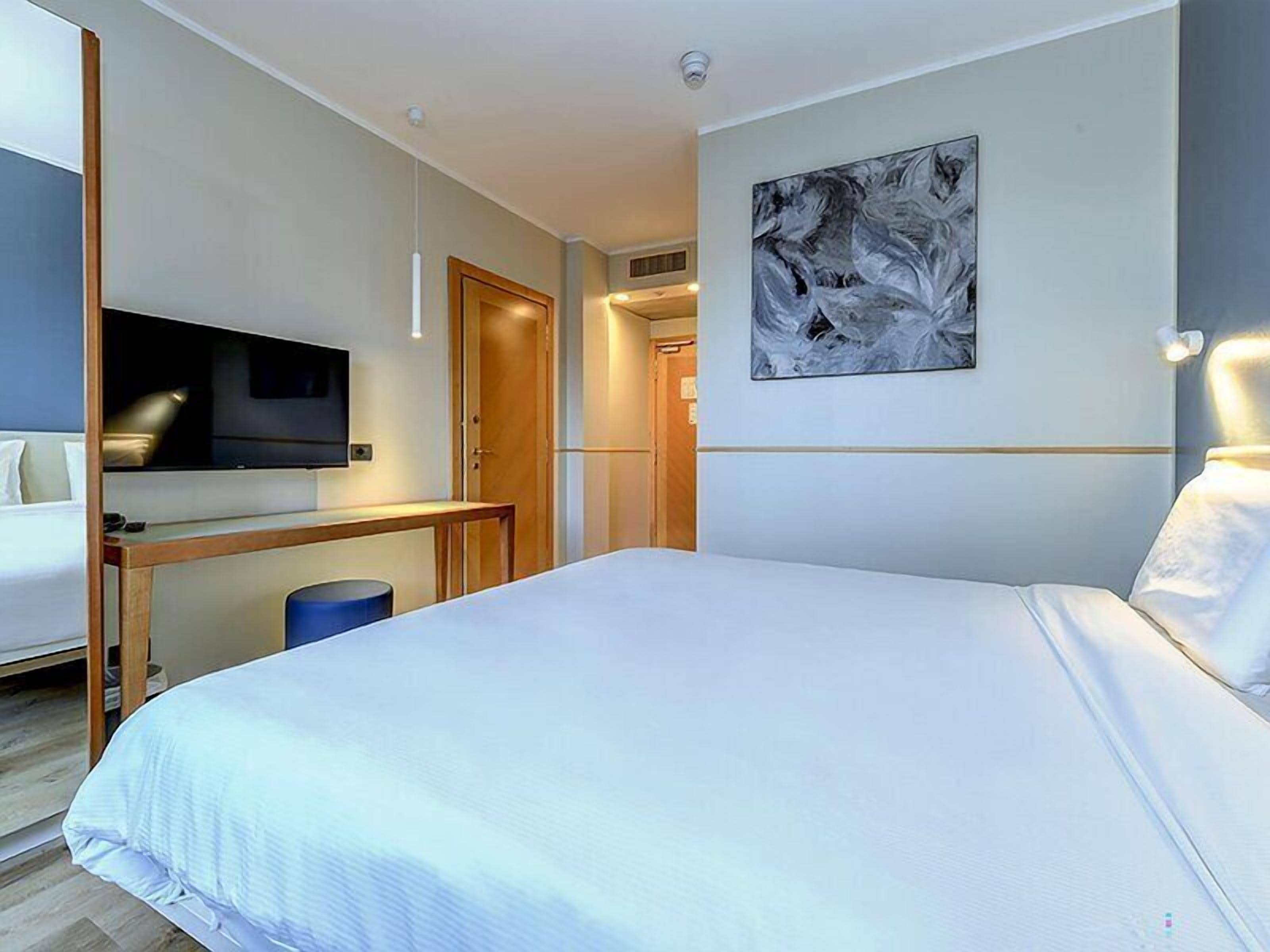 A modern hotel room with a large bed, a flat-screen TV mounted on the wall, a wooden desk, and a blue trash bin.