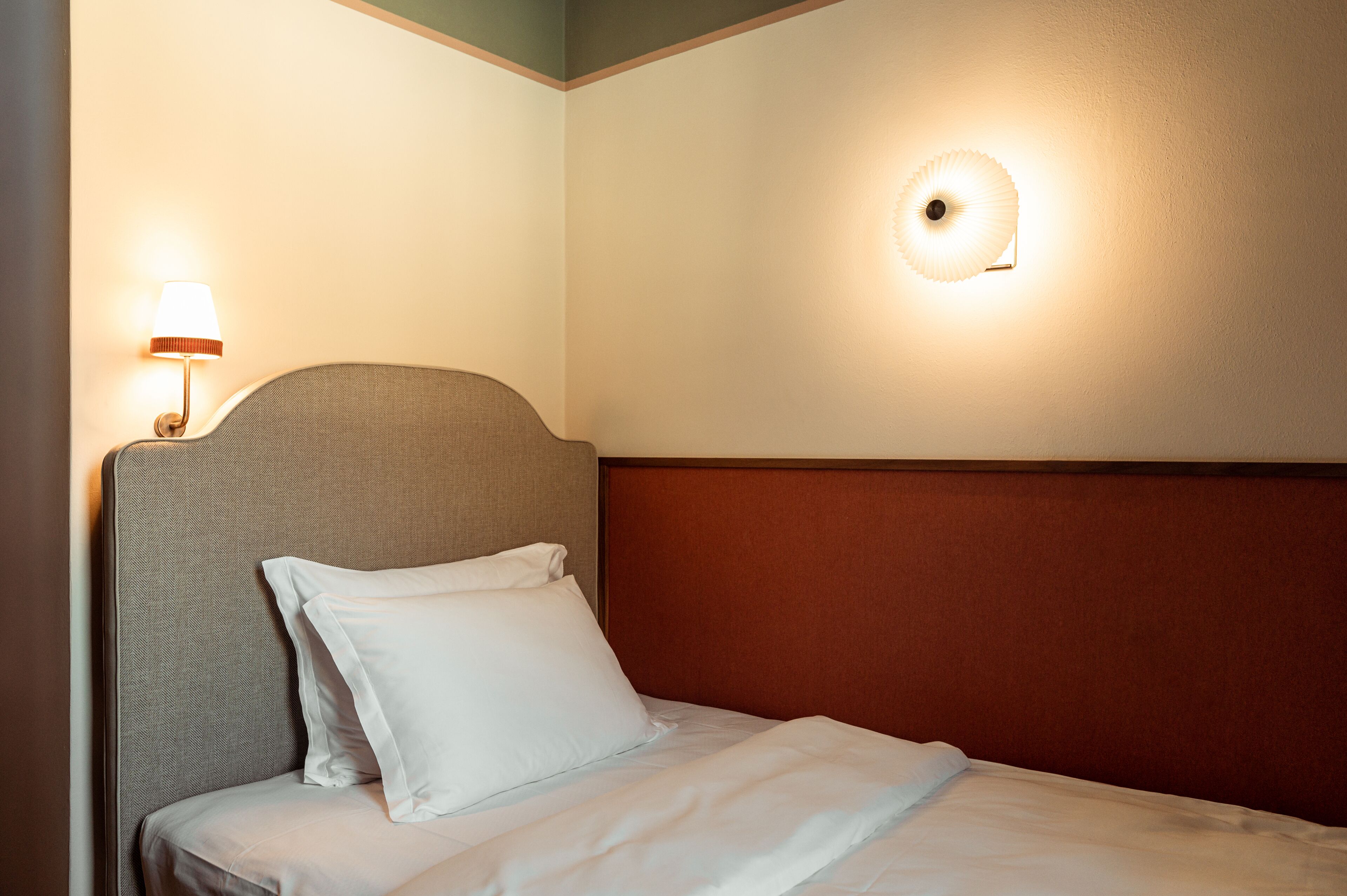 A hotel room with a bed, a nightstand, a lamp, and a wall-mounted light fixture.