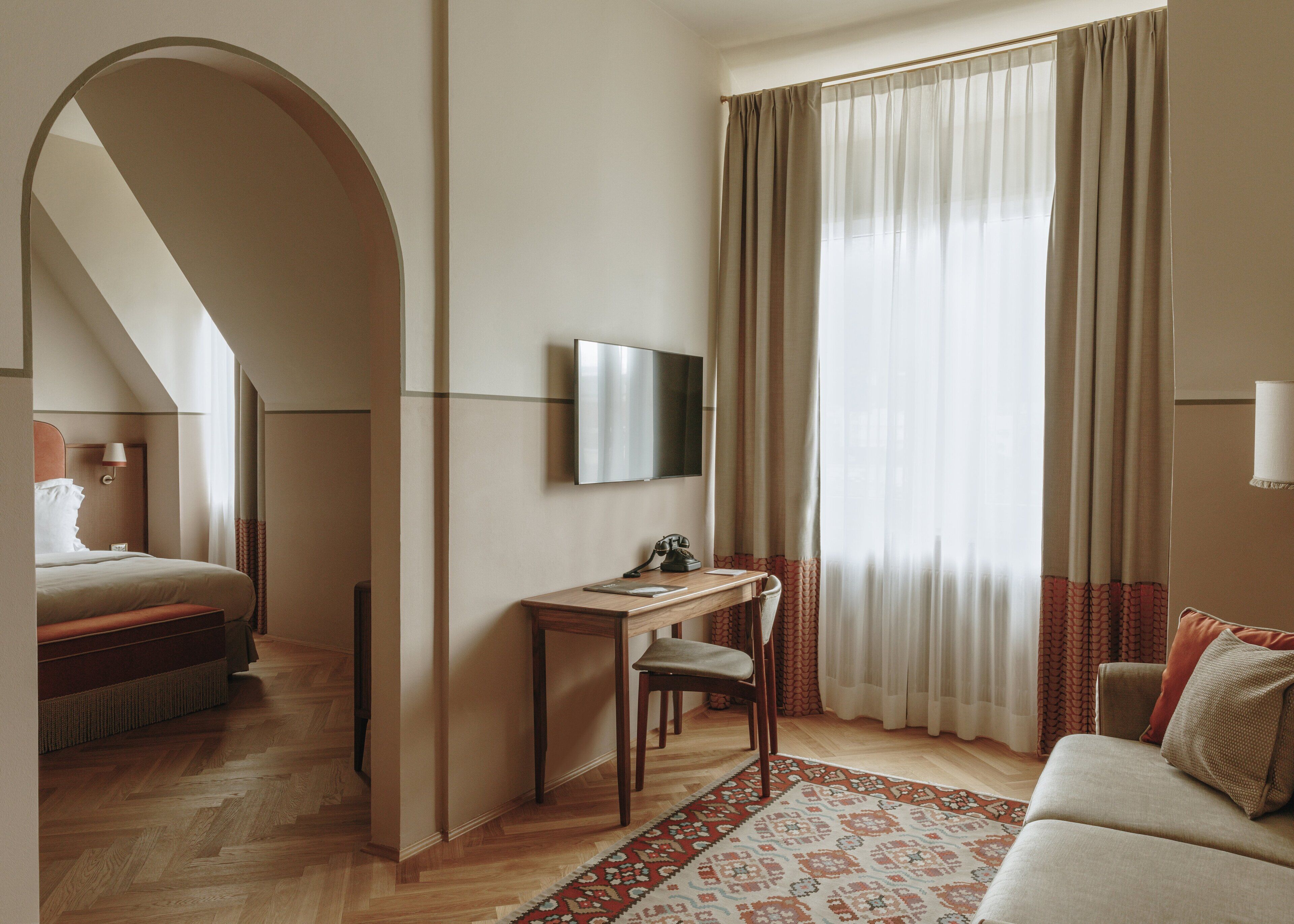 A hotel room with a bed, a desk with a chair, a TV, and a large window with curtains.