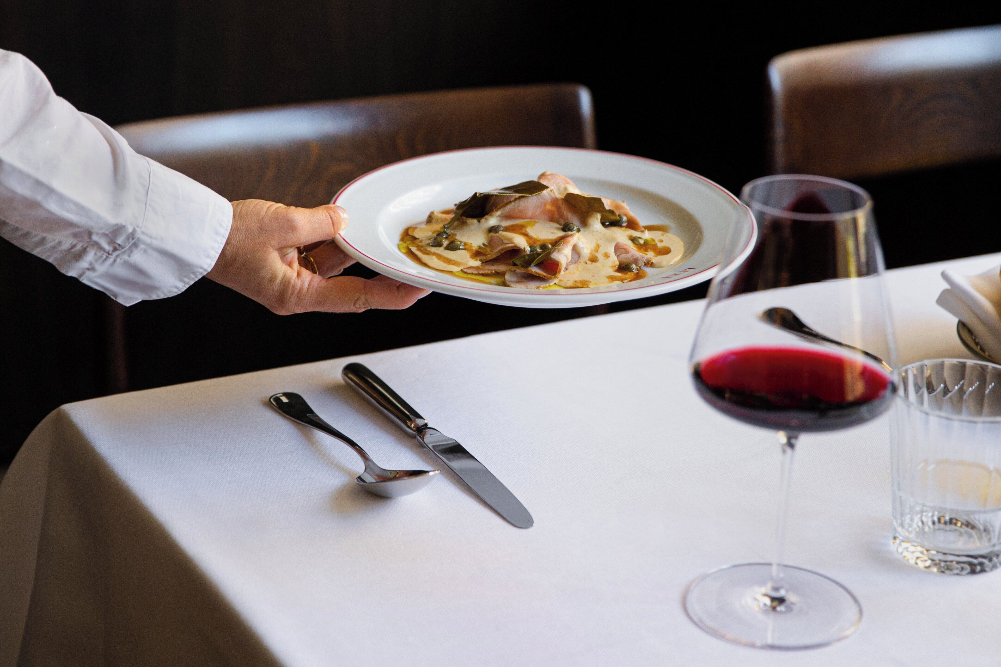 Eine Person hält einen Teller mit Essen an einem Tisch, auf dem Besteck und ein Glas Rotwein stehen.