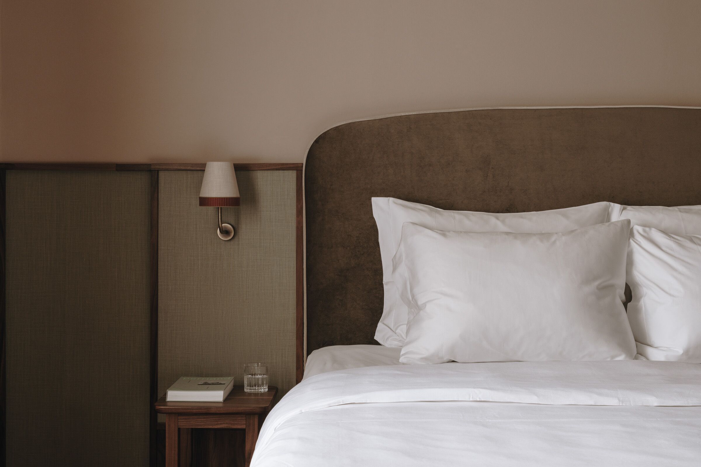 Un lit bien fait, avec du linge de lit blanc, une lampe de chevet et une petite table sur laquelle se trouvent un livre et un verre.
