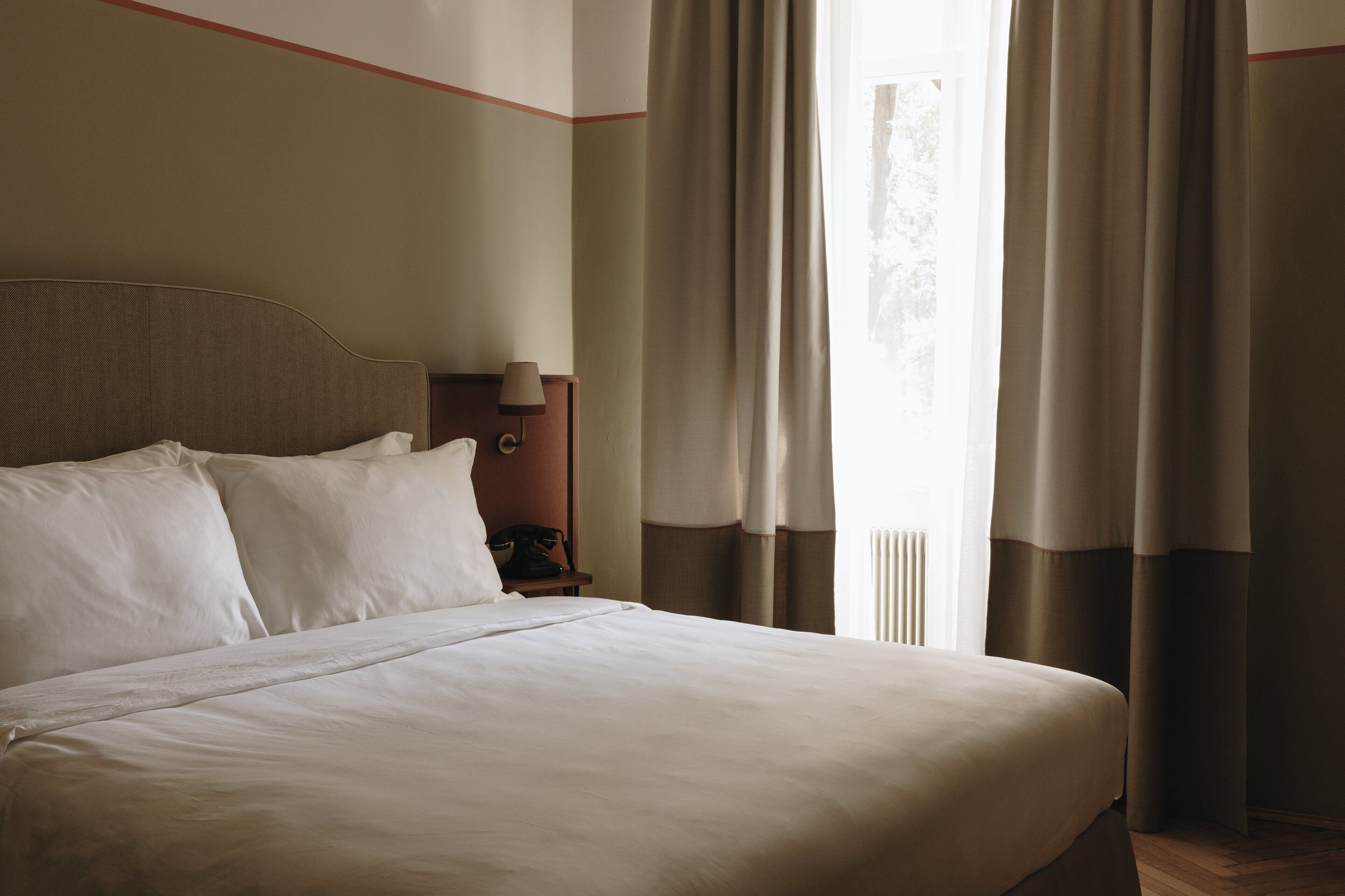 Un llit ben fet a una habitació d'hotel, amb capçalera, una taula de nit amb una làmpada i una finestra amb cortines.