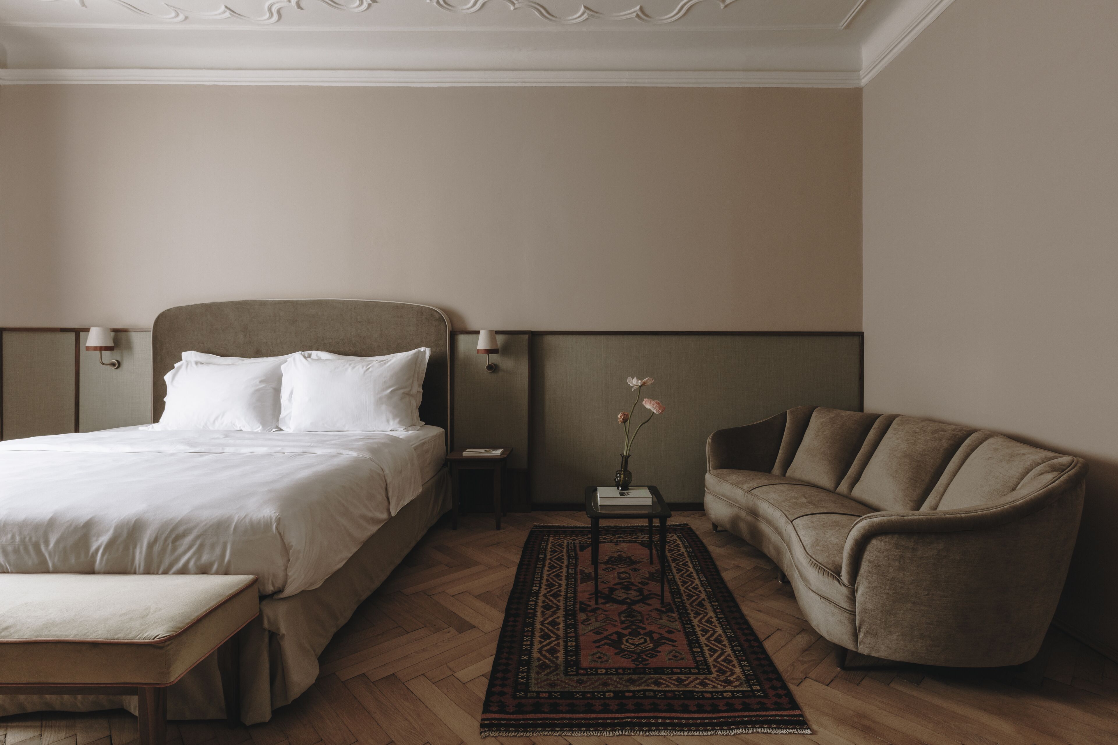 A hotel room with a bed, a nightstand, a vase with flowers, a sofa, and a bench.
