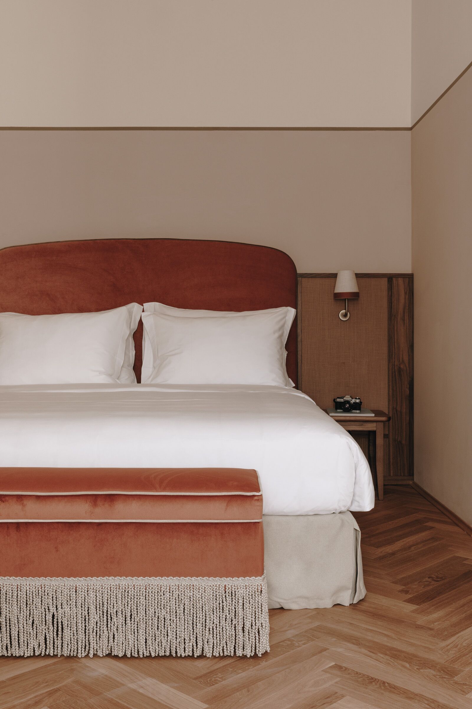 Un lit bien fait, avec une tête de lit rouge, une lampe de chevet et un appareil photo posé sur une petite table à côté.