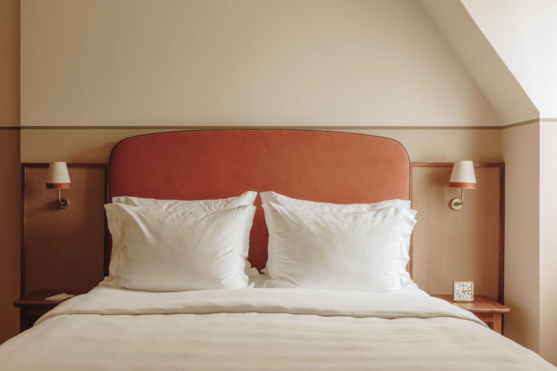 Una cama bien hecha con sábanas blancas y un cabecero rojo, flanqueada por dos mesitas de noche con lámparas.