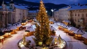 Una plaza cubierta de nieve con un gran árbol navideño decorado en el centro, rodeada de edificios con tejados nevados.