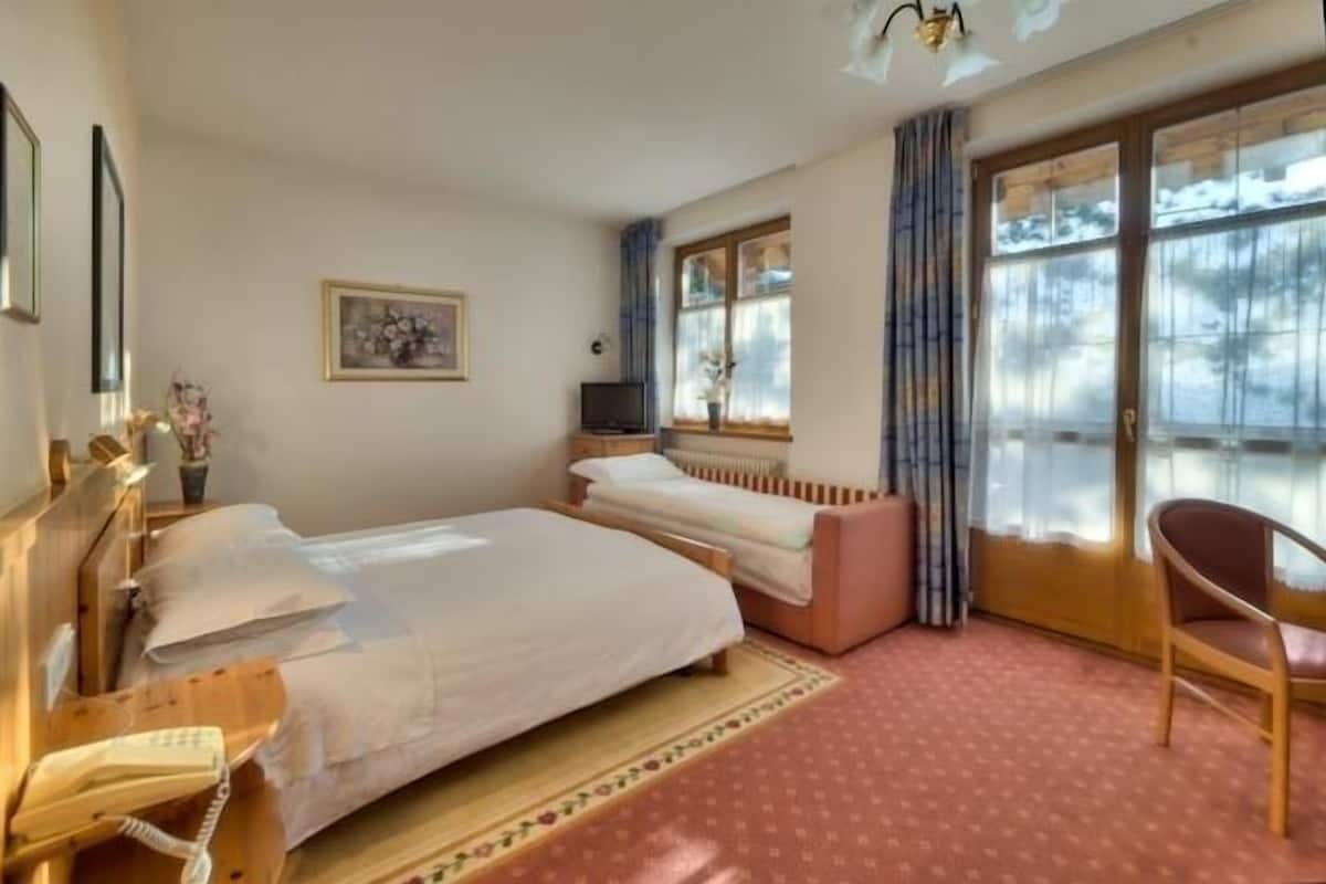 A hotel room with two beds, a chair, a desk with a telephone, a radiator, and a window with curtains.