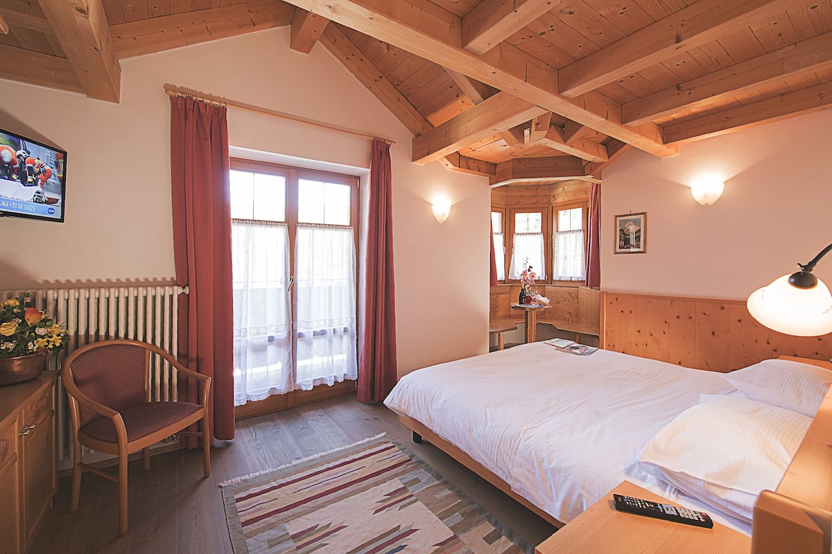 A bedroom with a wooden ceiling, a bed, a chair, a nightstand, a lamp, a window with curtains, and a radiator.