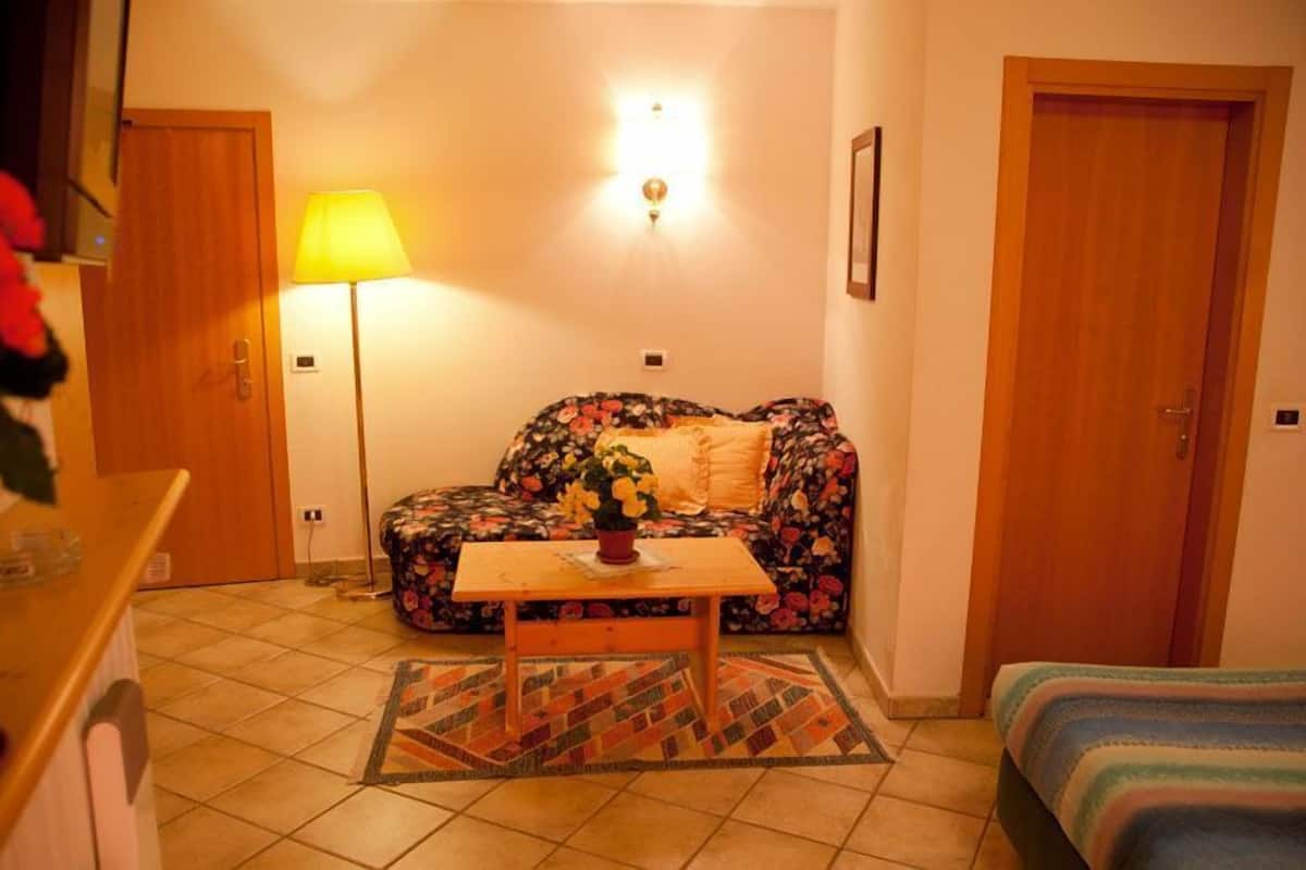 A cozy room with a floral sofa, a small wooden table with a vase, a lamp, and a bed.