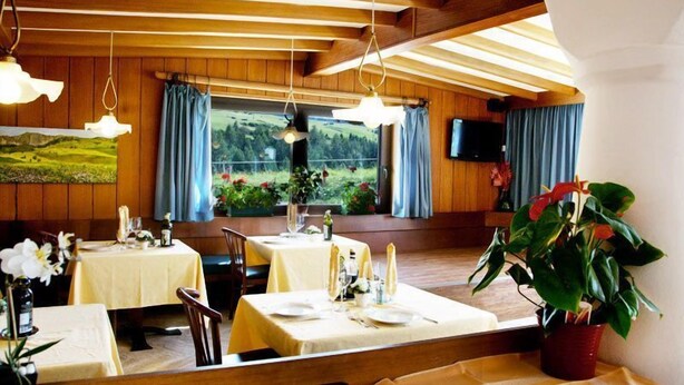 A dining area with tables set for two, wooden beams on the ceiling, and a large window with a view of greenery. - Piccolo Hotel Sciliar (Castelrotto)