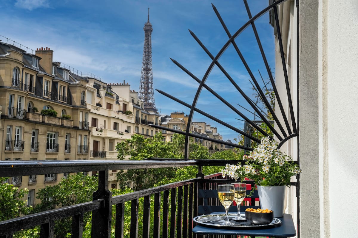 A balcony with a table set for two, overlooking a cityscape with historic buildings and the Eiffel Tower in the background.
