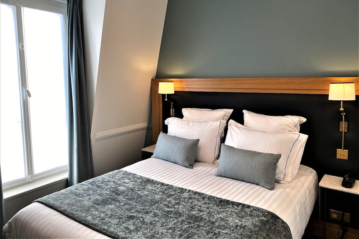 A hotel room with a bed, two bedside lamps, a window with curtains, and a dark wall.