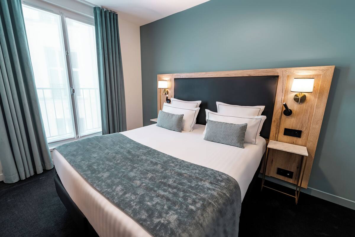 A hotel room with a large bed, two bedside tables, a nightstand, a window with curtains, and a wall-mounted lamp.
