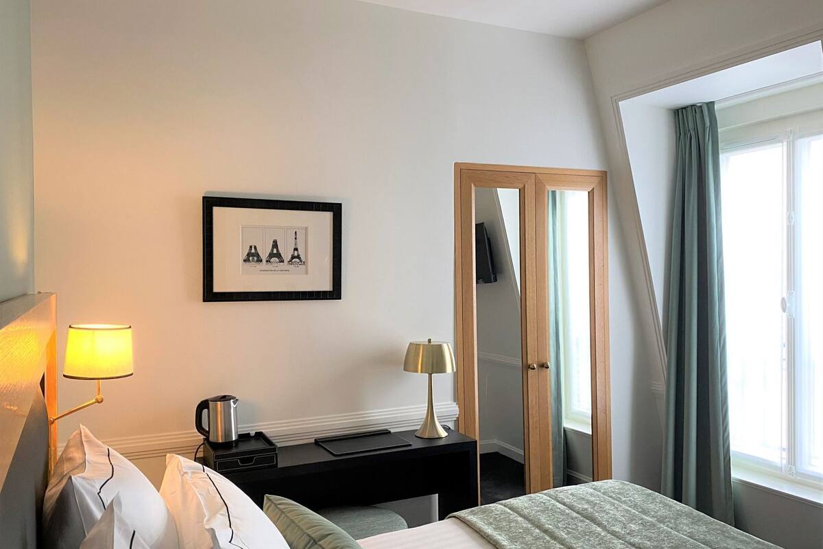 A hotel room with a bed, bedside lamps, a framed picture of the Eiffel Tower, a desk with a kettle, and a window with curtains.