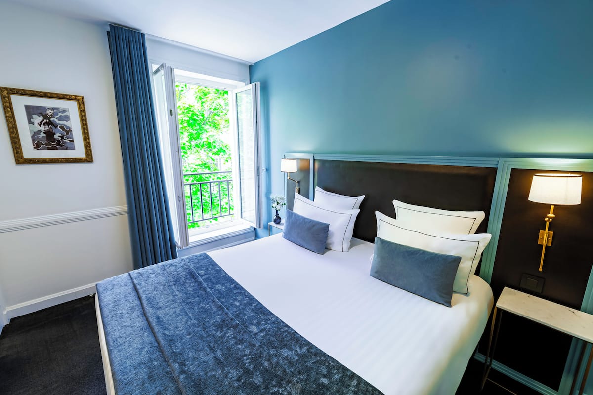 A hotel room with a large bed, a nightstand with a lamp, a window with a view of greenery, and a framed picture on the wall.
