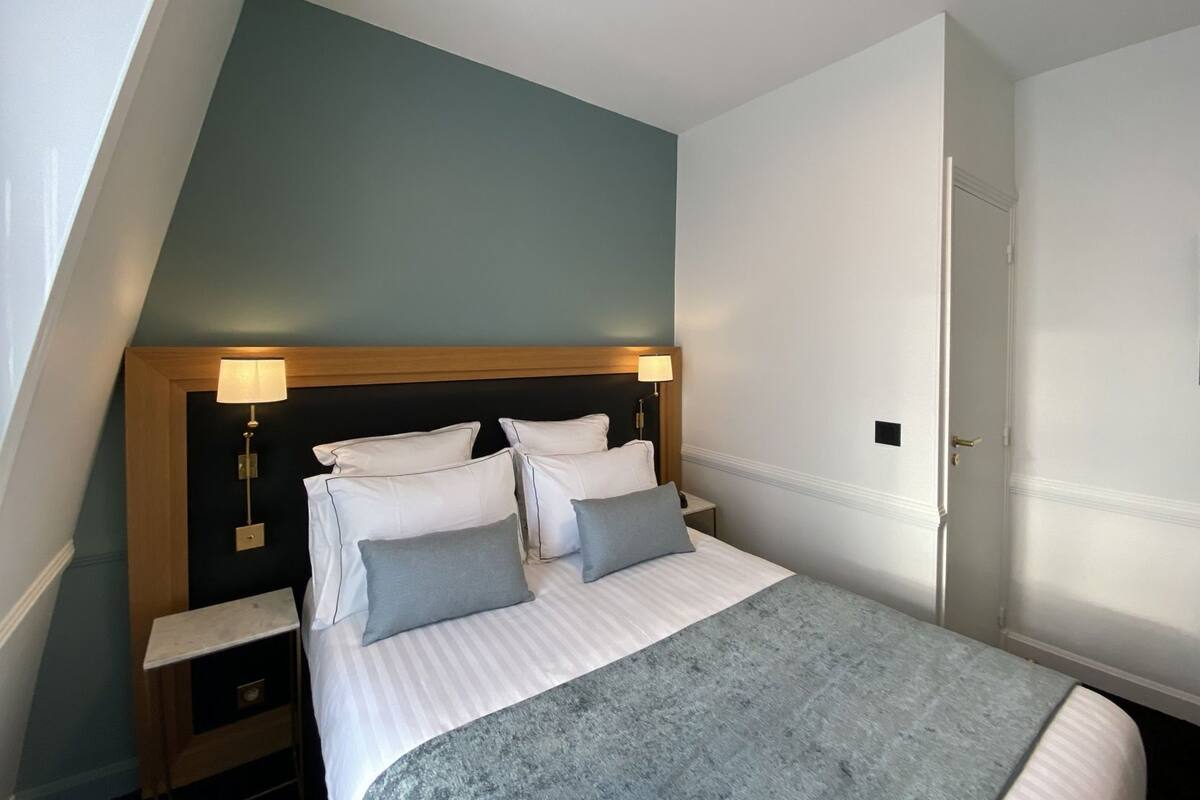 A bedroom with a sloped ceiling, a bed with white bedding and two pillows, a bedside table with a lamp, and a door to the right.