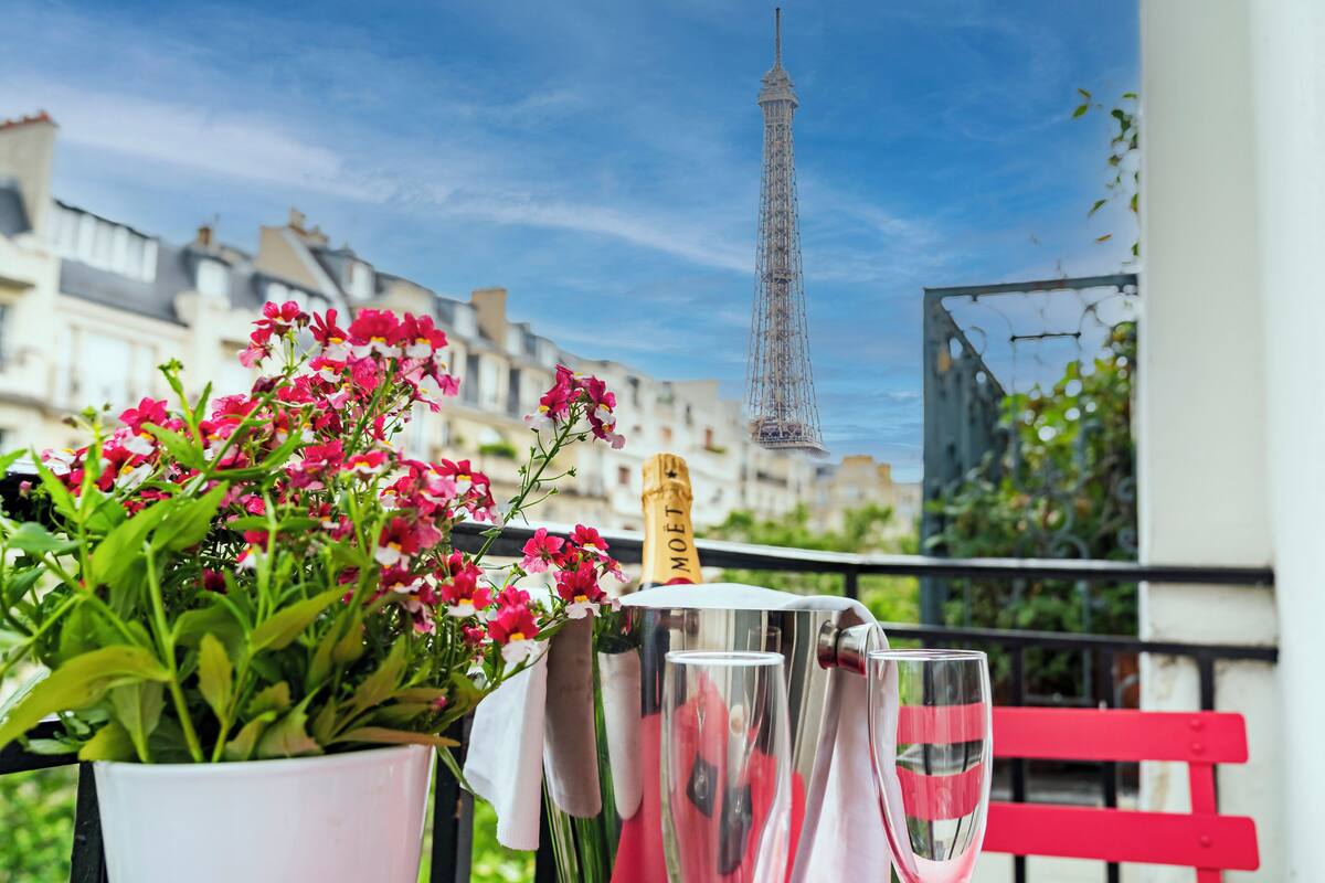A balcony with a potted plant, a bottle of champagne, and champagne glasses. The Eiffel Tower is visible in the background.