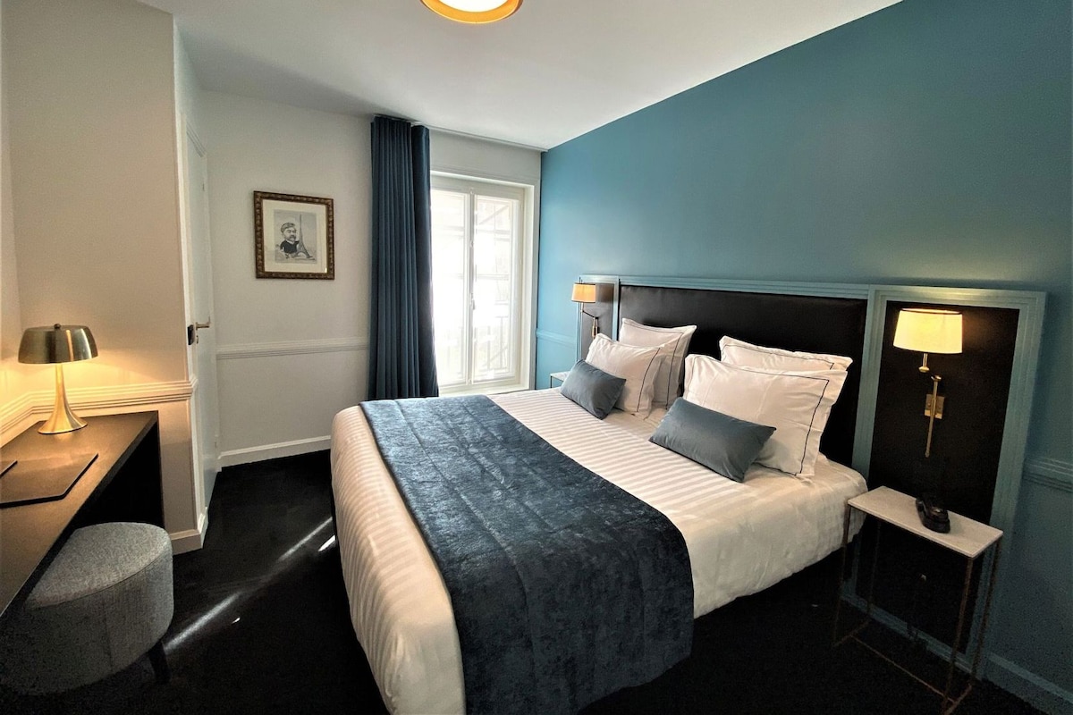 Quarto de hotel com cama, mesas de cabeceira, escrivaninha, cadeira e janela com cortinas.