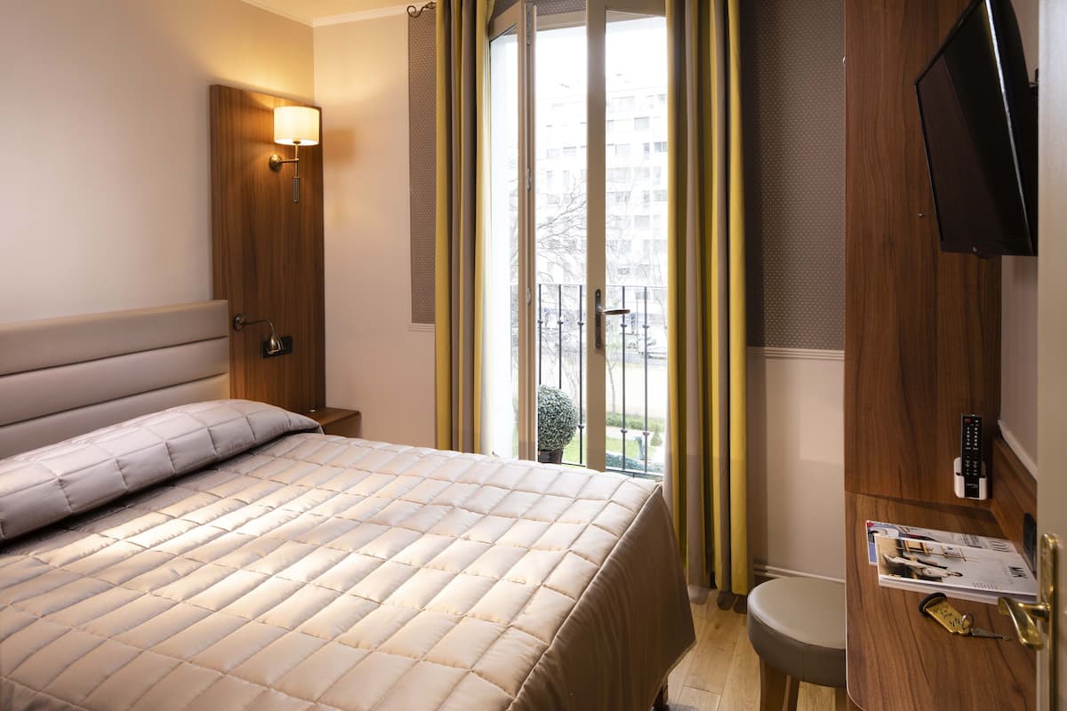 A hotel room with a bed, a bedside table, a lamp, a TV, and a window with curtains.