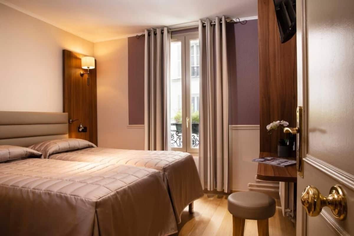 A hotel room with two beds, a small table, a bench, a wall-mounted lamp, and a door with a mirror and a potted plant.