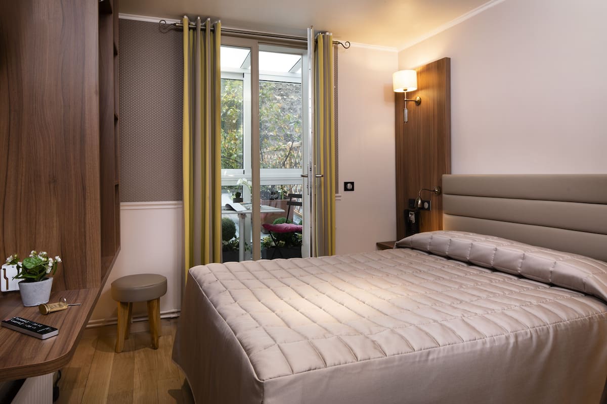 A bedroom with a bed, a desk, a chair, a small stool, and a window with curtains.