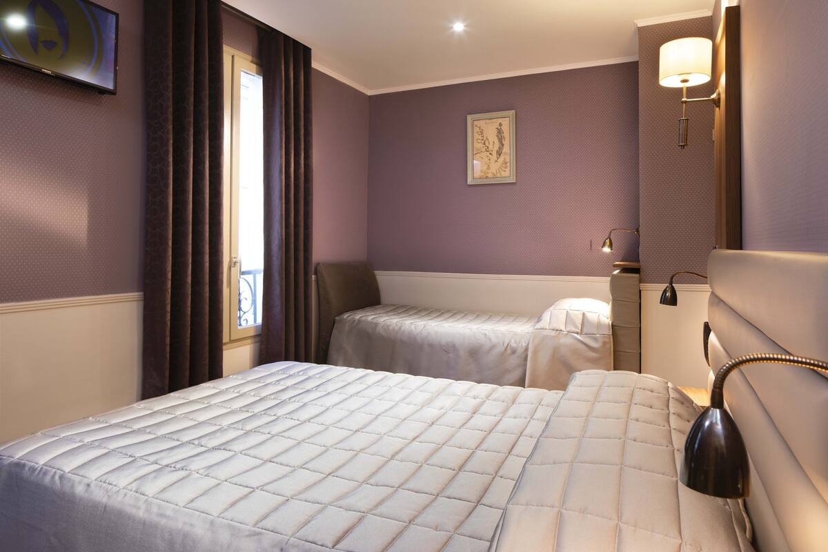 A hotel room with two beds, a window with curtains, a wall-mounted lamp, and a painting.